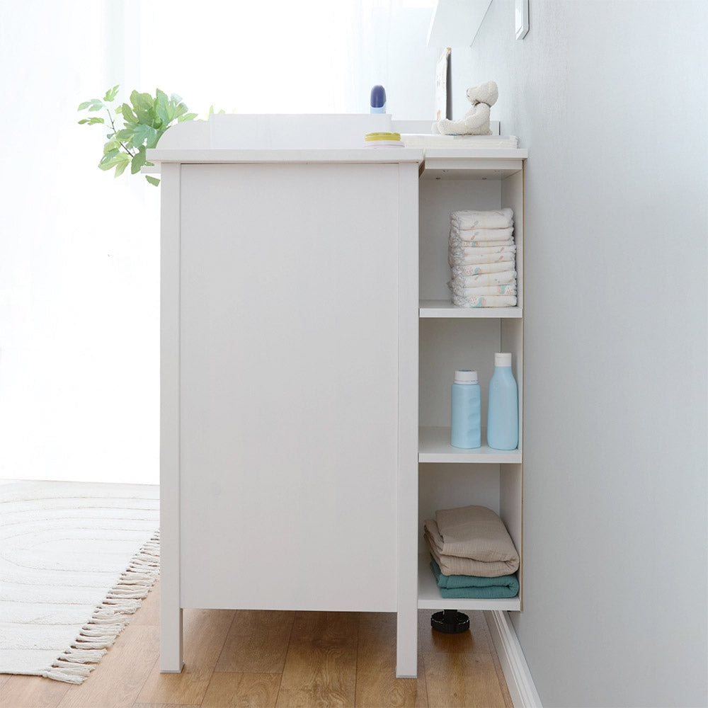 Seitliche Ansicht eines umgebauten Wickeltisches, bestehend aus einer weißen Hemnes Kommode mit einem passenden Wickelaufsatz. Rechts befindet sich ein seitlich angebautes offenes Stauraumregal mit drei Fächern, die Windeln, Pflegeprodukte und gefaltete Textilien enthalten.