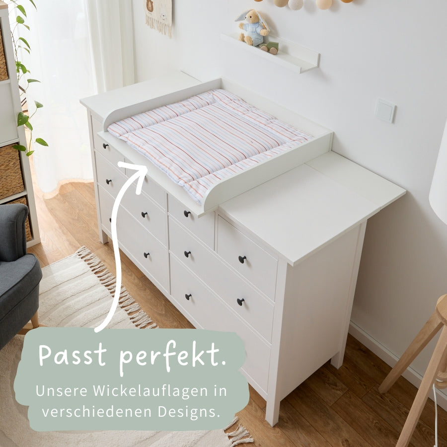 Eine IKEA Hemnes Kommode, mit 8 Schubladen, auf der sich ein weißer Wickelaufsatz und eine passgenaue Wickelauflage mit Streifen befindet.