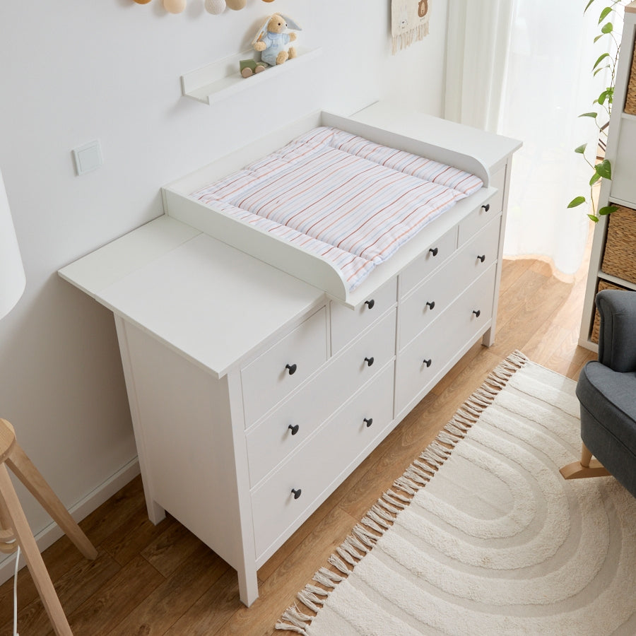 Eine große IKEA Hemnes Kommode, auf der unser Wickelaufsatz, inklusive einer Wickelauflage mit dezentem Streifen-Muster, montiert ist. Um die Kommode herum ist eine schöne Kindereinrichtung zu sehen - mit einem Schaukelstuhl, einem Kallax mit Körben, flauschigm Teppich, einem Häschen und einer natürlichen Lampe.